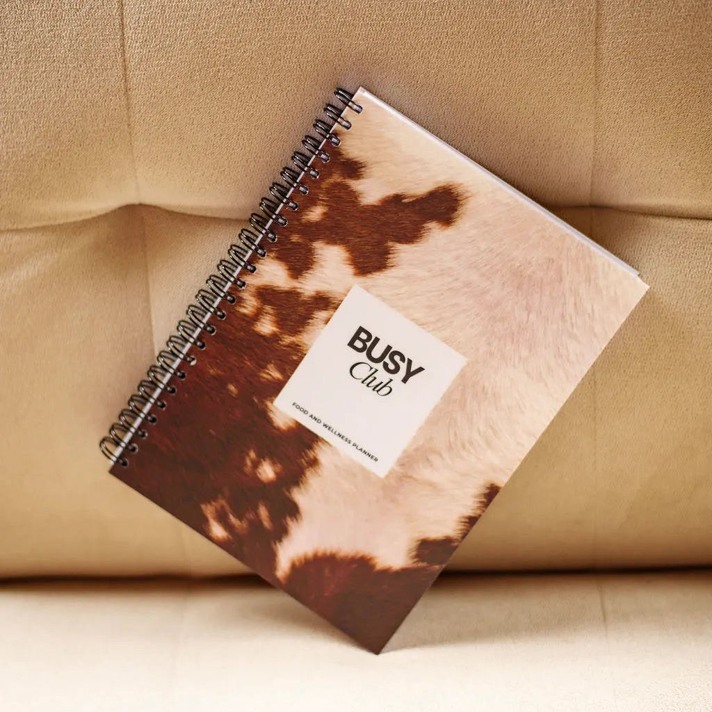 Notebook with cow print cover and 'BUSY Club' label on a beige couch