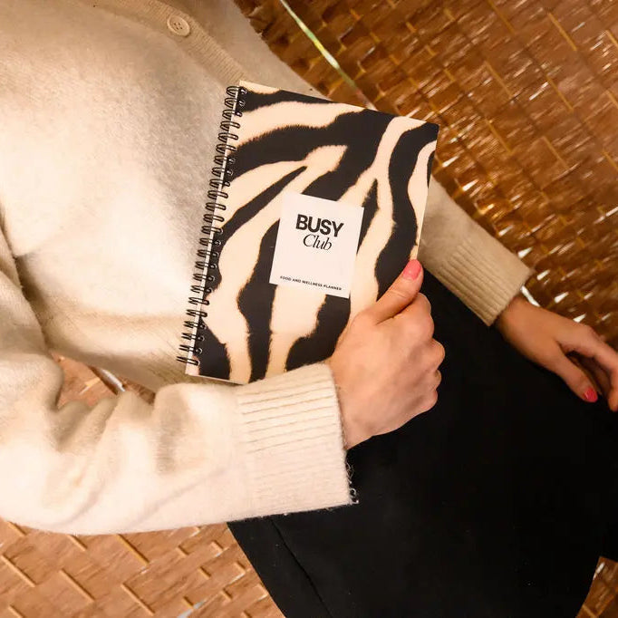 Person holding a zebra print notebook with 'BUSY Club' branding against a woven surface.