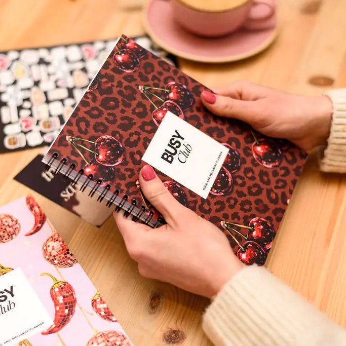 Person holding a leopard print notebook with 'BUSY Club' branding on a wooden table.