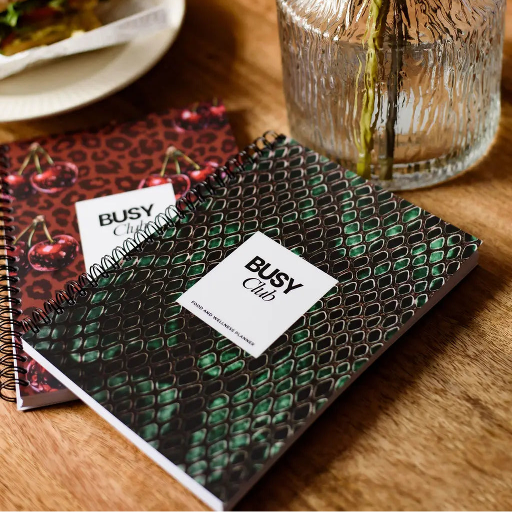 Two notebooks with 'BUSY Club' branding on a wooden table with a vase and food in the background.
